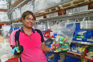 A woman in a pink uniform sorts colourful highlighters in a warehouse with organised office supplies.